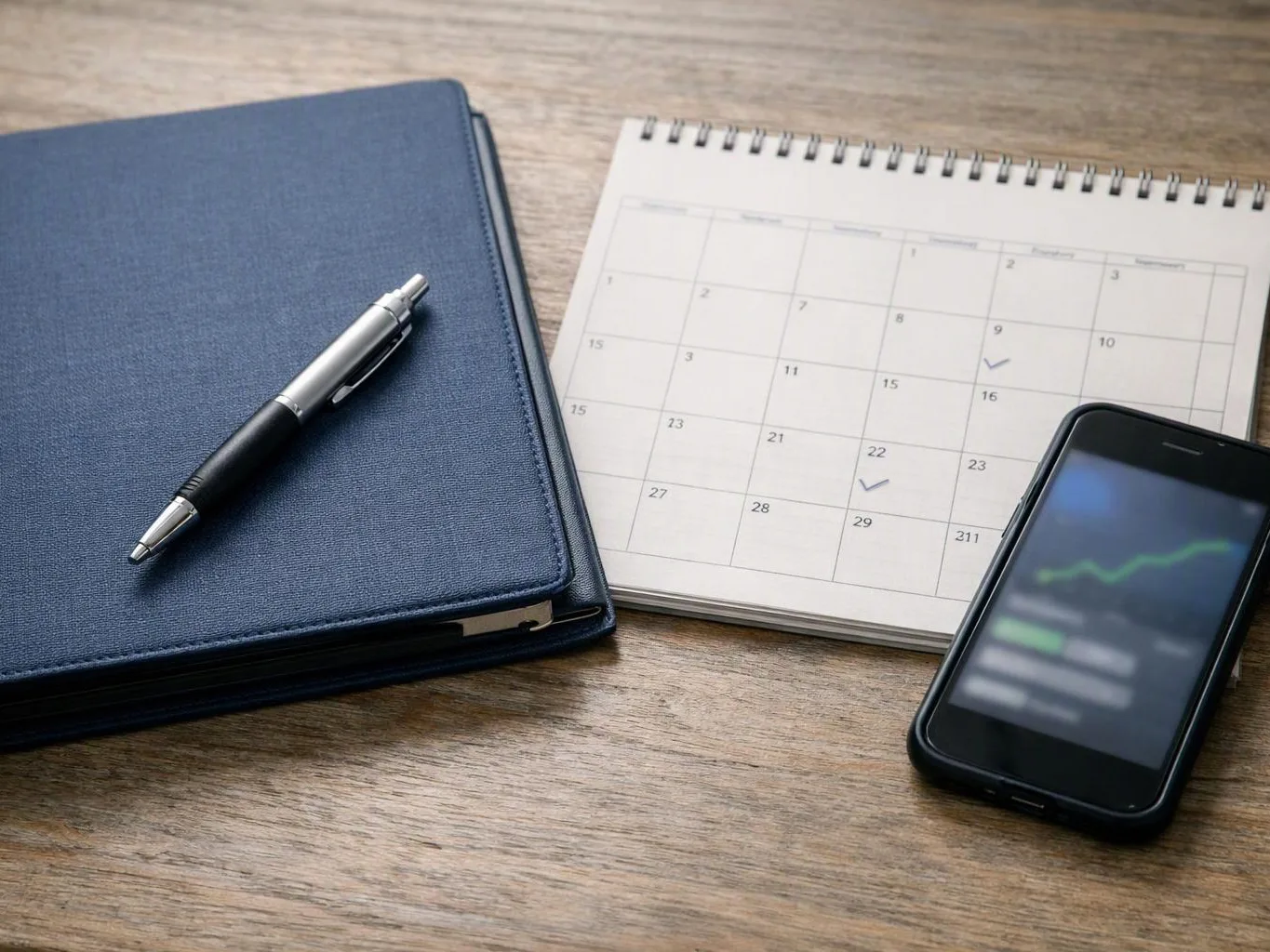 A blue notebook with a pen sits beside a spiral calendar and a smartphone on a wooden desk