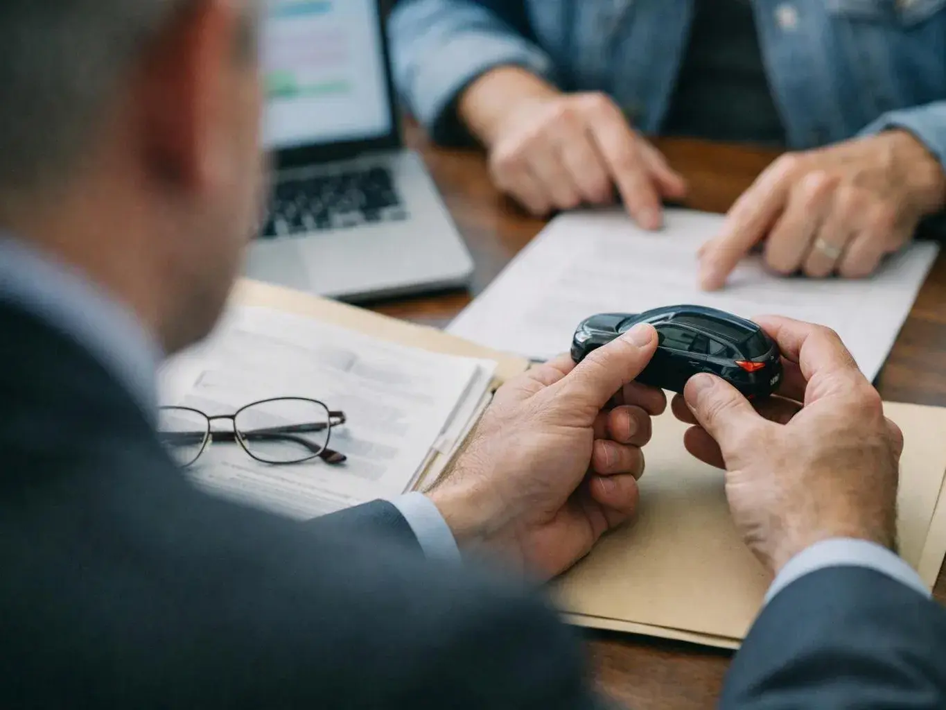 A man holds a small black toy car over papers during a meeting at a wooden table