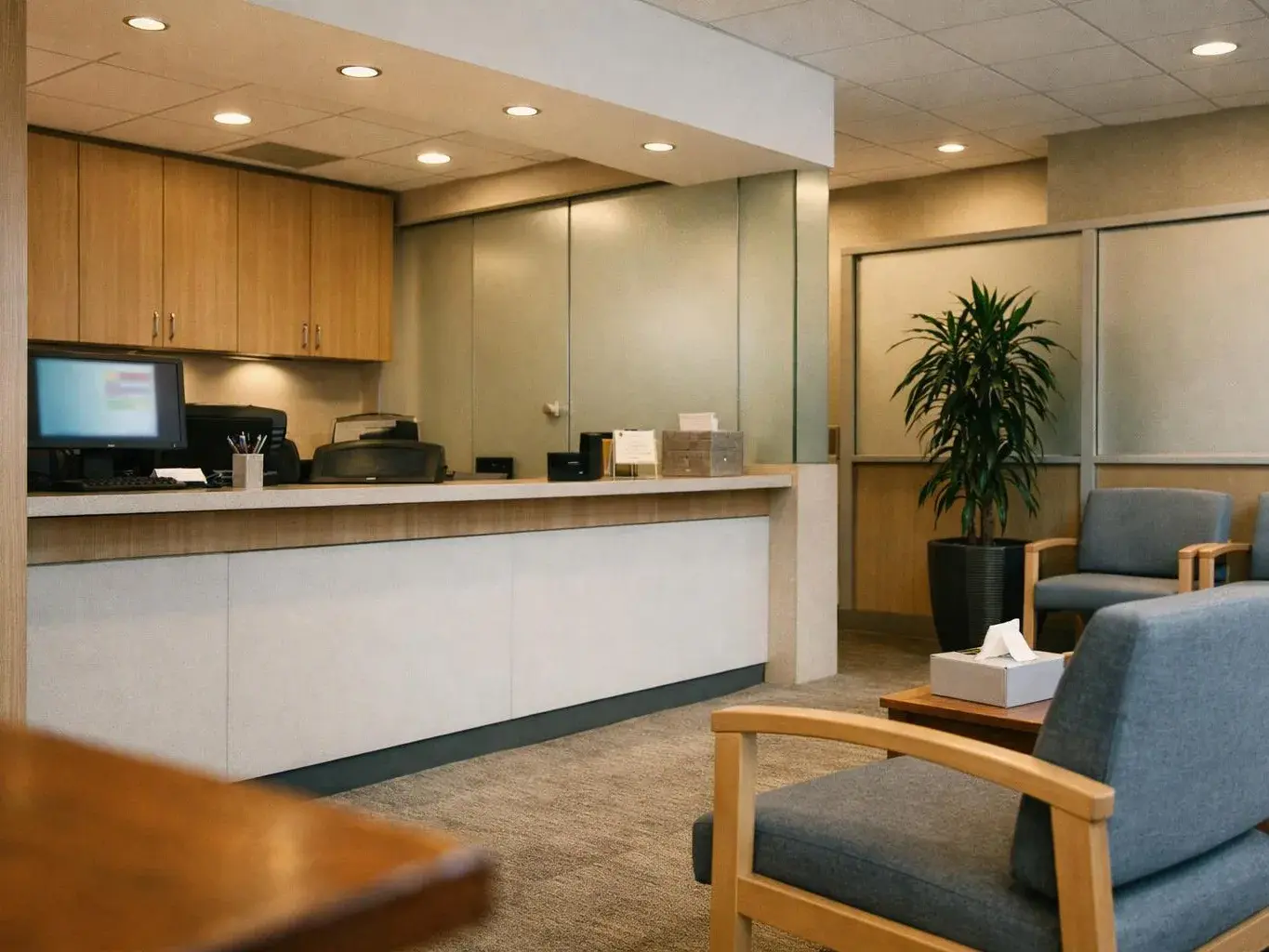 A quiet office reception area with a counter, chairs, a potted plant, and recessed ceiling lights