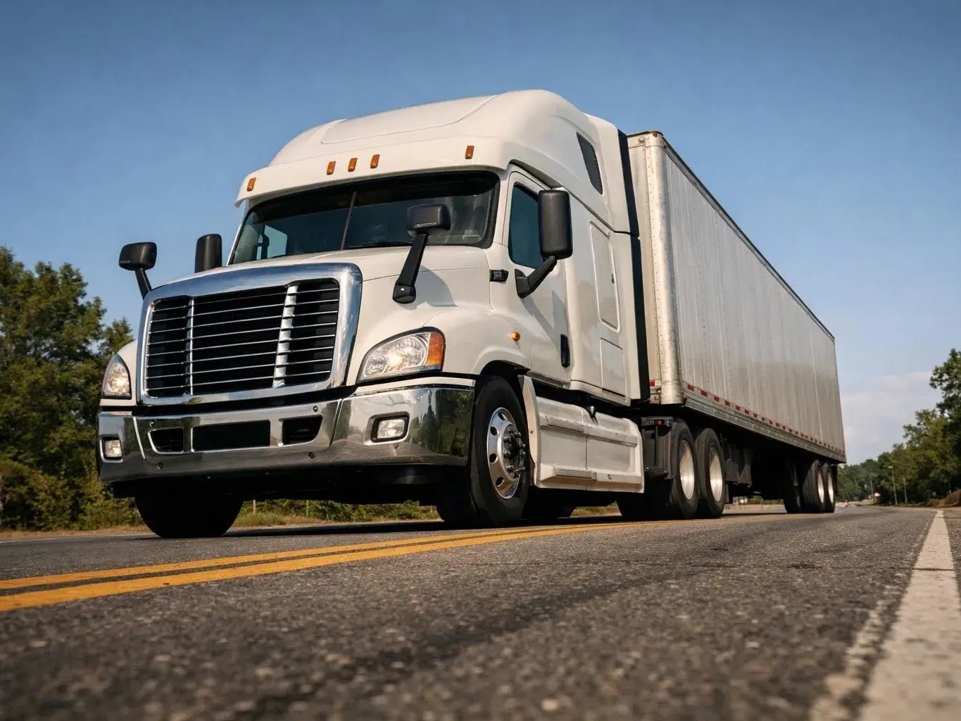 A white semi truck pulls a long trailer down a two-lane road under a clear blue sky